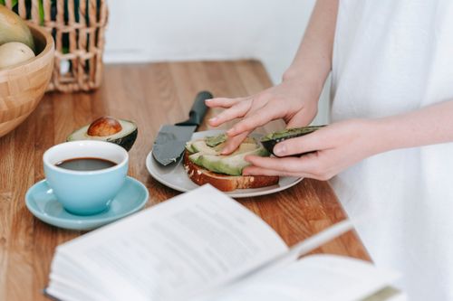 Free Crop anonymous female in white t shirt preparing toast with sliced avocado and cup of coffee for breakfast at wooden table Stock Photo