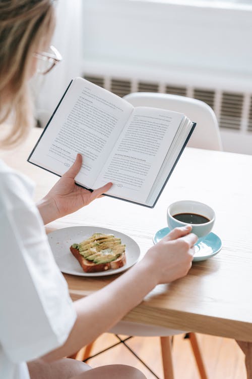Free Crop unrecognizable female in white t shirt sitting at table with cup of black coffee and avocado toast while reading book Stock Photo