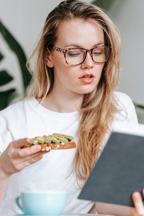 Free Young female in white clothes and glasses sitting at table and eating toast while reading book with interest Stock Photo