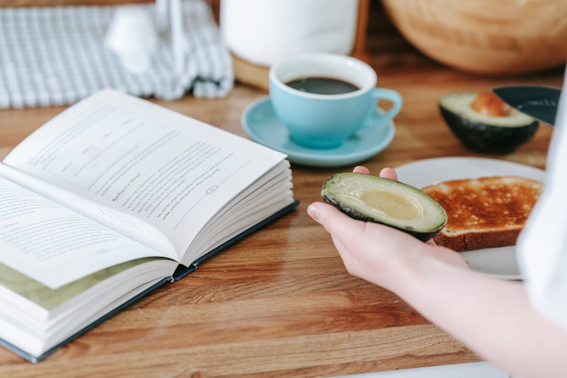 Free Crop anonymous person sitting at table and reading book while having coffee and making toast with avocado for breakfast Stock Photo