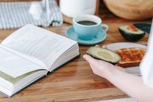 Crop anonymous person sitting at table and reading book while having coffee and making toast with avocado for breakfast