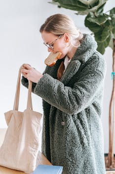 Young female in warm jacket with glasses standing near table with paper bag and holding piece of bread in mouth