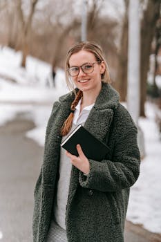 Modern happy female in stylish coat with book looking at camera while standing on snowy road near forest on blurred background