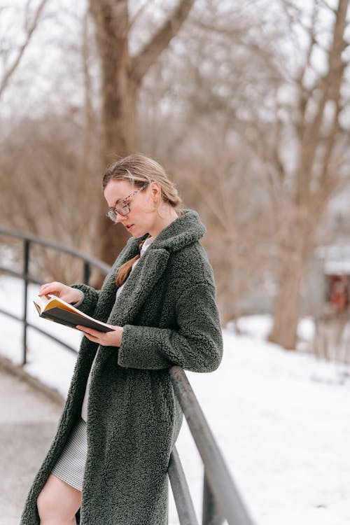 Side view of focused female in warm clothes and eyeglasses reading book while standing near railing in city snowy park