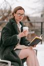 Modern calm female in warm coat and with shopper bag sitting on wooden bench holding book and takeaway cup of coffee while resting in winter park