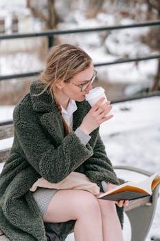 Young female in warm clothes with takeaway hot drink sitting on bench and reading book in winter park