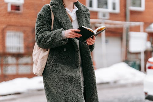 Crop unrecognizable woman in coat with textile bag reading textbook while studying on city street