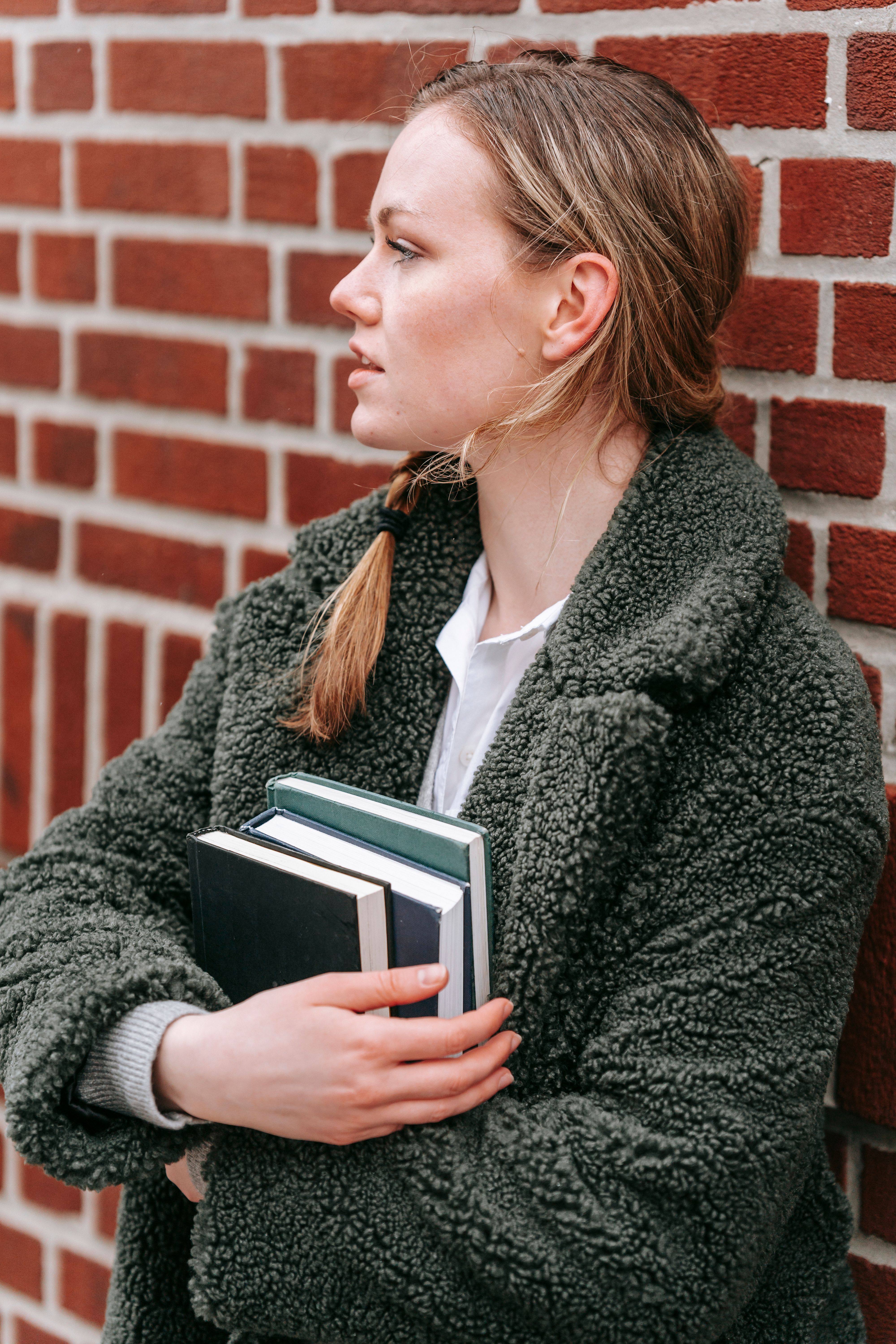Contemplative student with textbooks against brick wall · Free Stock Photo