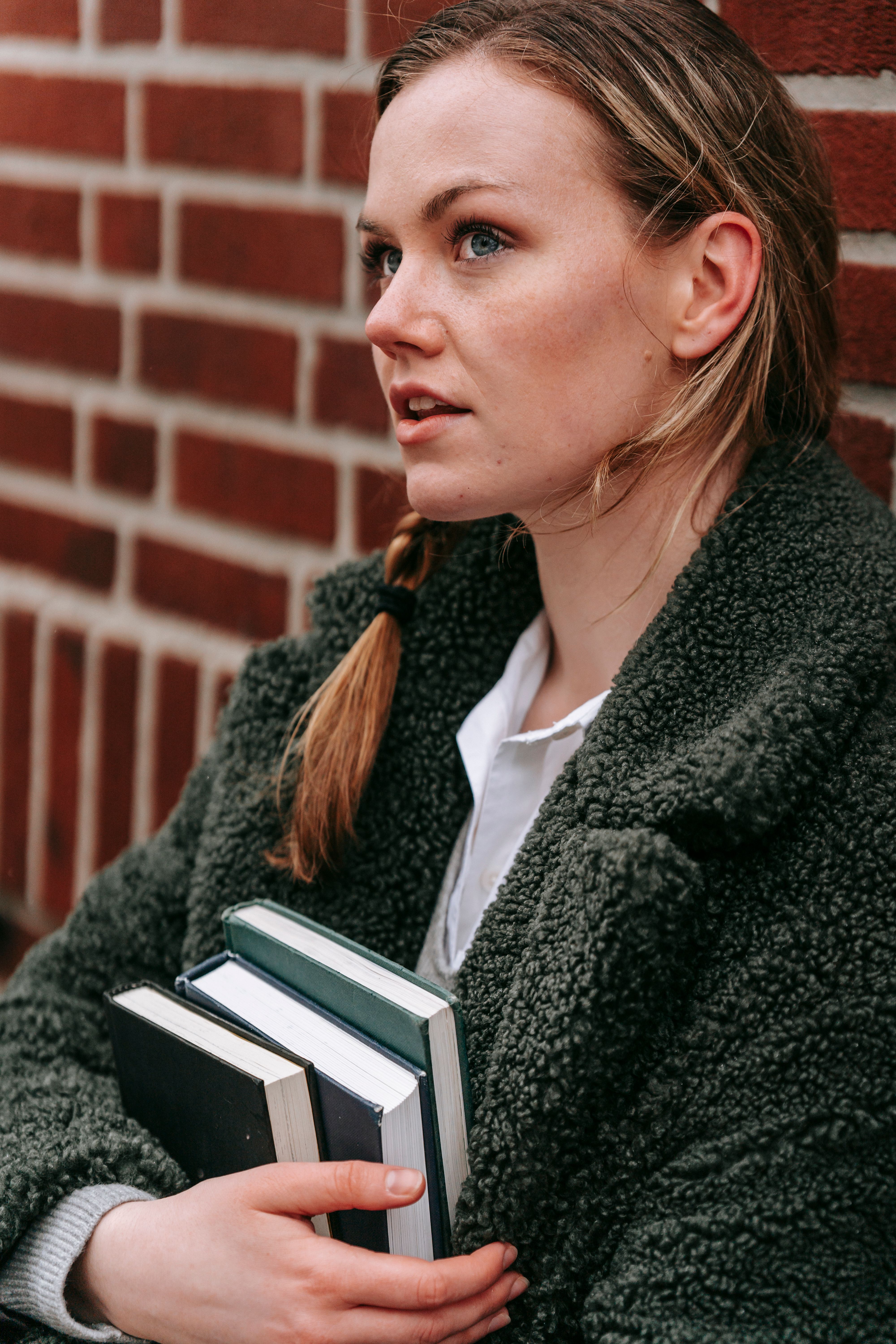 Thoughtful young woman in three-quarter profile gazing into the distance, clutching a stack of books against a brick wall