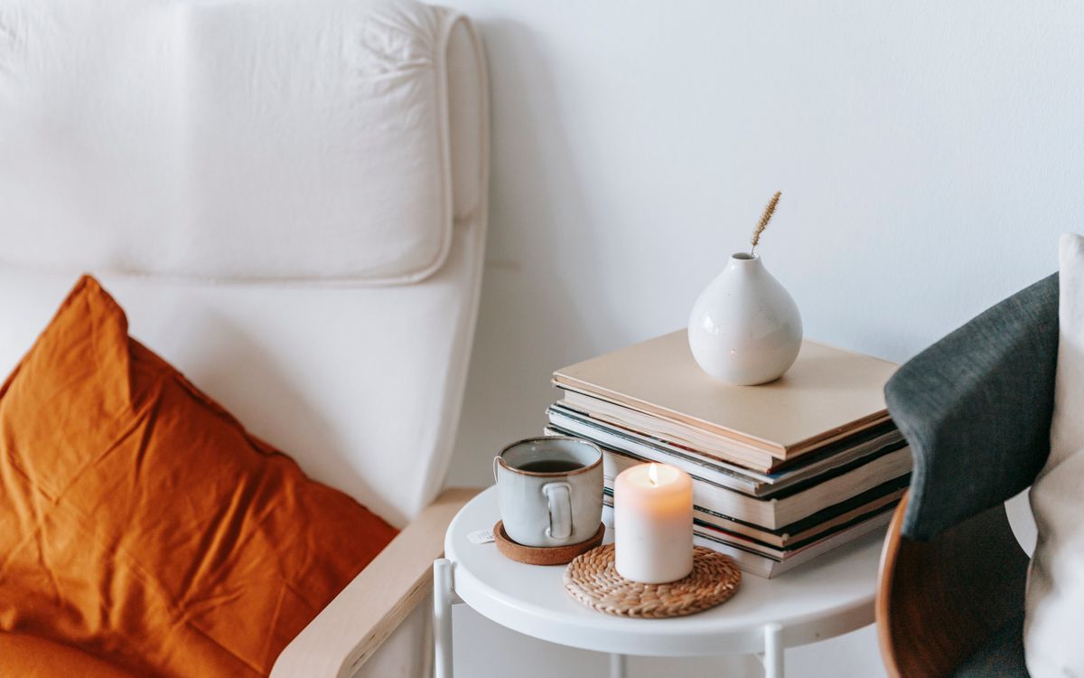 Cup of tea and flaming candle against heap of books with dry spike in vase between armchairs at home on white background