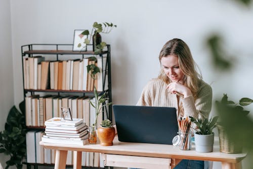 Interested young female remote employee surfing internet on netbook at table with potted plants against bookshelves in house on light background