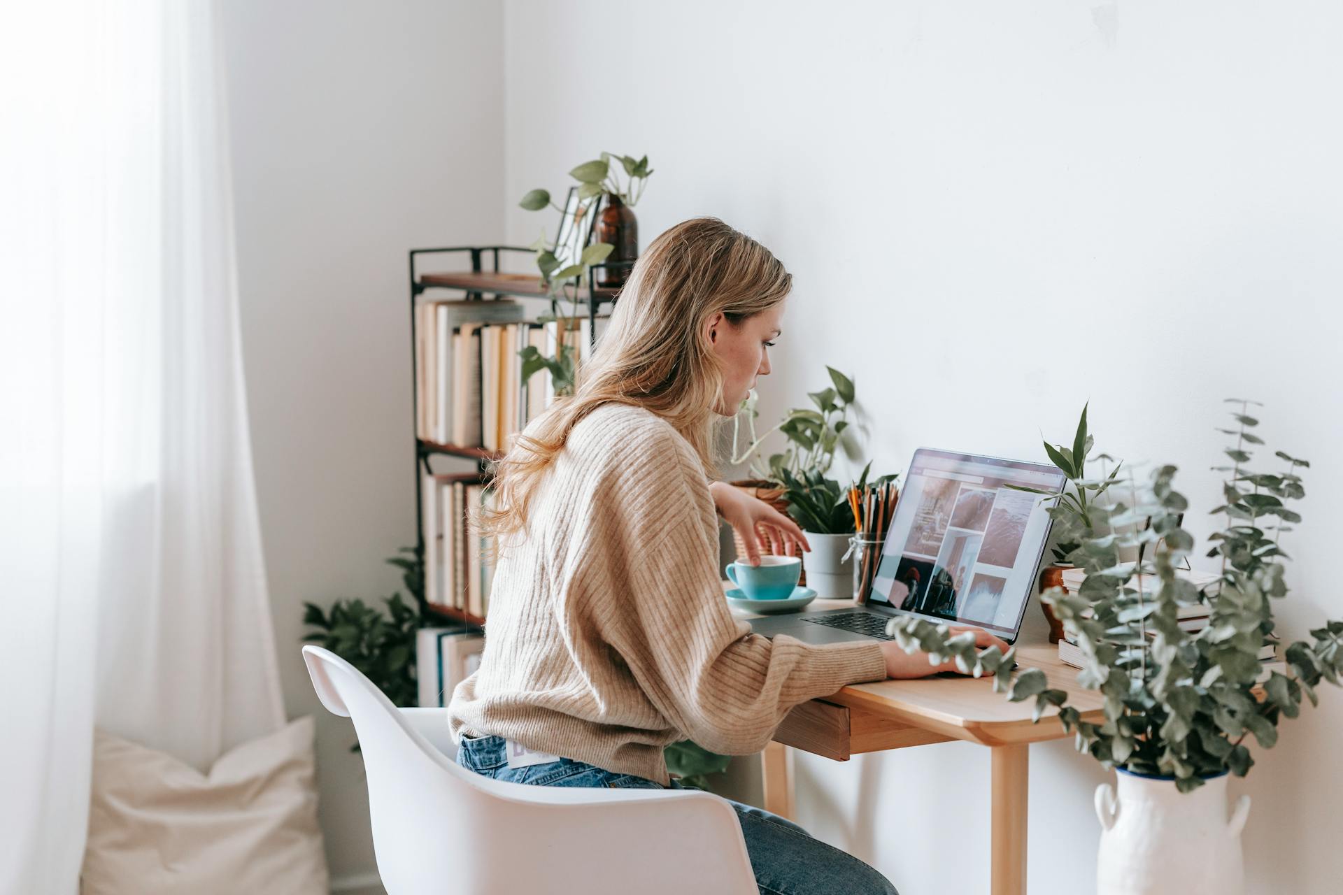 Side view of attentive female remote employee with cup of tea watching photo gallery on netbook screen at desk in house