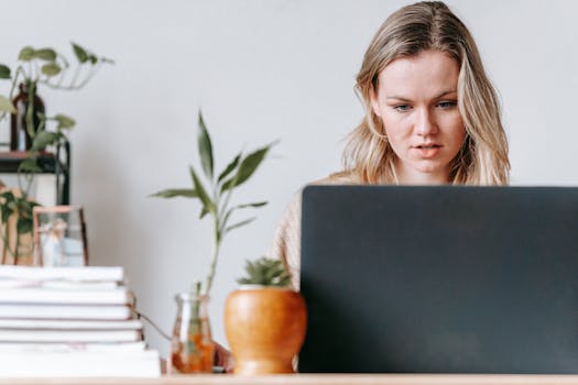 Young woman concentrating on her laptop in a home office setting, surrounded by houseplants and books.