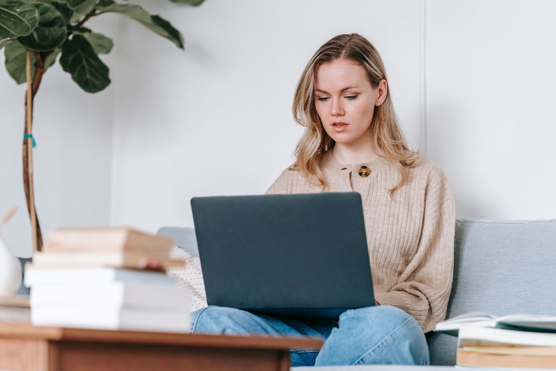 Focused young woman using laptop for study at home, surrounded by books and cozy decor.