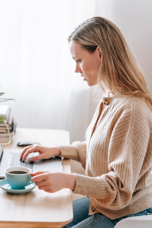 Side view of attentive female remote worker with hot drink browsing internet on netbook while sitting at table in house