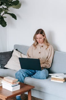 Young serious female in casual clothes using netbook while working remotely at table with books in lounge