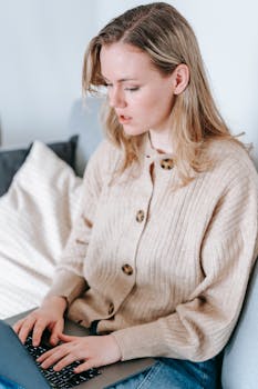 A young woman in a cardigan focused on her laptop indoors, conveying a casual work environment.