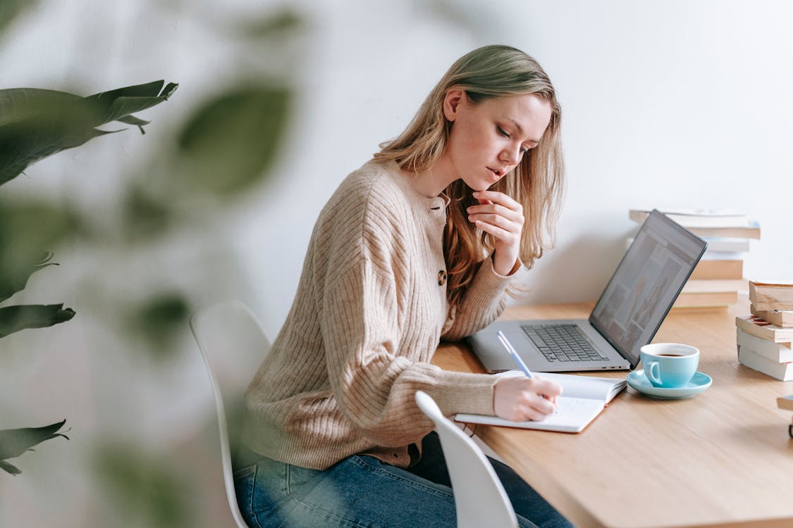 Free Pensive woman taking notes at table with laptop and cup of coffee Stock Photo