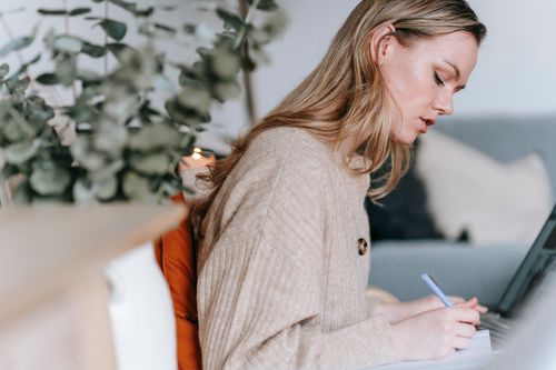Free Side view of crop gentle serious female taking notes in planner near netbook at table on blurred background Stock Photo