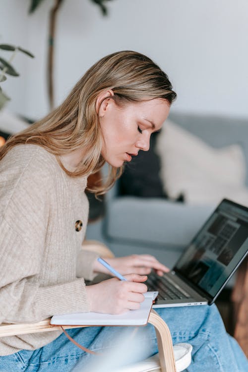 Side view of focused young female taking notes in diary while browsing netbook on blurred background