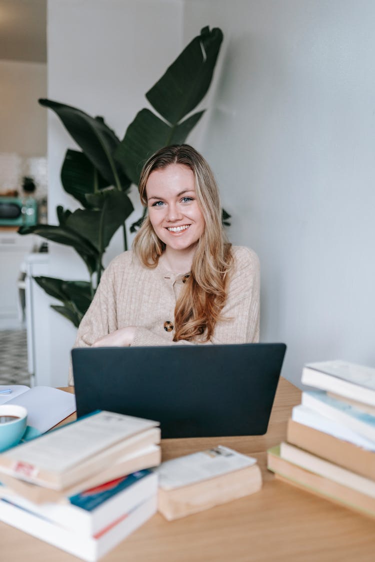 Cheerful Student At Table With Laptop And Heap Of Books