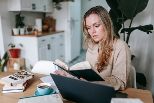 Young focused female studying planner near netbook and cup of hot aromatic coffee on blurred background of kitchen interior