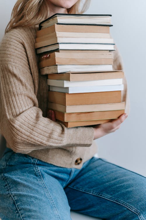 Crop unrecognizable female student in casual clothes holding stack of books against light wall in daytime
