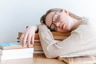 Exhausted female student in eyeglasses and casual sweater sitting at table and sleeping on books on light room