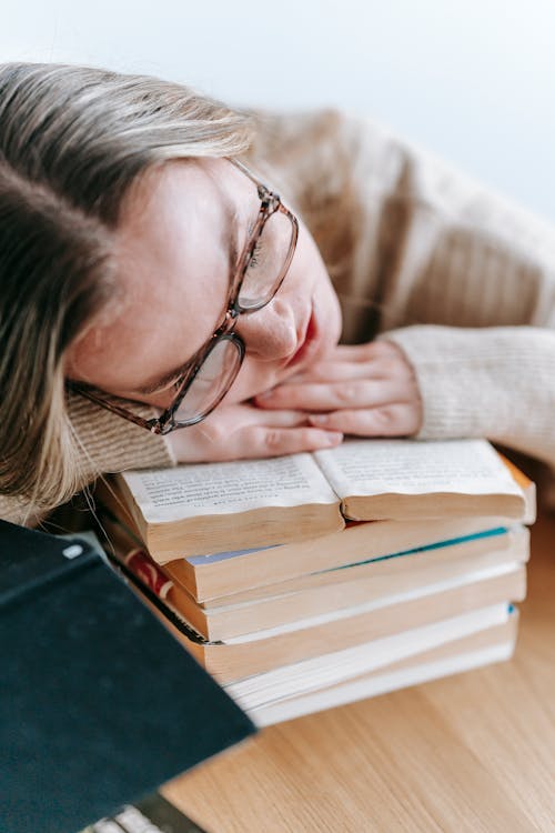 Free Tired intelligent female in eyeglasses sitting at table and sleeping on pile of books while studying Stock Photo