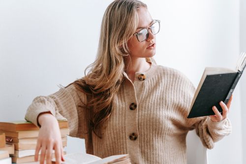 Free Young female in casual clothes with eyeglasses reading book while sitting at table near white wall Stock Photo
