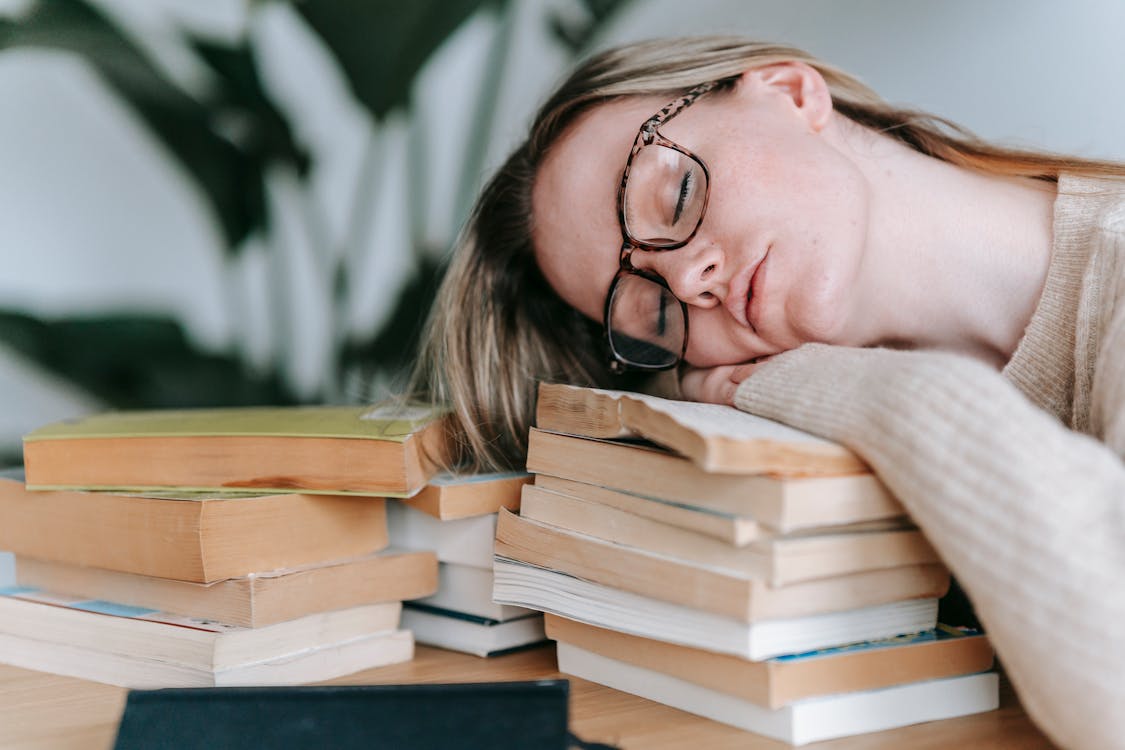 Free Tired female student with eyeglasses sitting at table and sleeping on pile of books on white background with plant Stock Photo