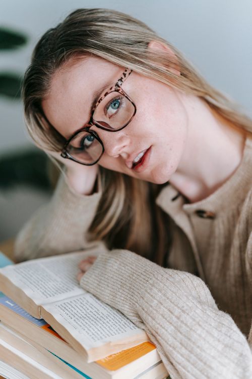 Free Young female in glasses with pile of books looking at camera while leaning on hand in light room Stock Photo