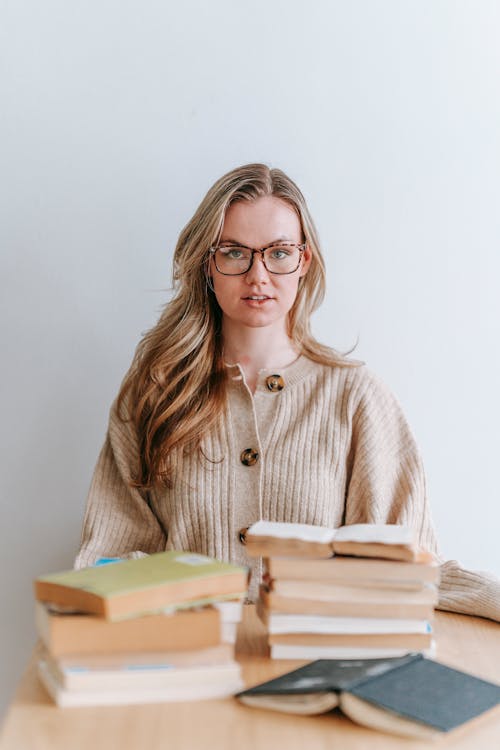 Free Smart female student in cardigan and eyeglasses sitting at desk with pile of books while looking at camera on white background Stock Photo