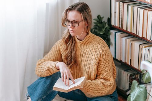 Free Pensive female student in casual clothes and eyeglasses sitting near bookshelf while reading book in light apartment Stock Photo