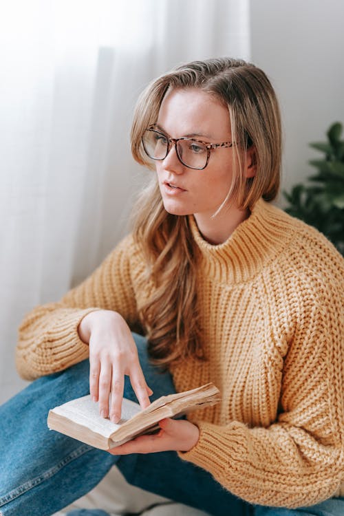 Free Pensive young female student in warm outfit and eyeglasses sitting with book while looking away in light white room Stock Photo