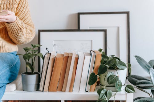 Crop female in casual clothes holding opened book and sitting on shelf with collection of books and plants and photo frames on white wall in light room