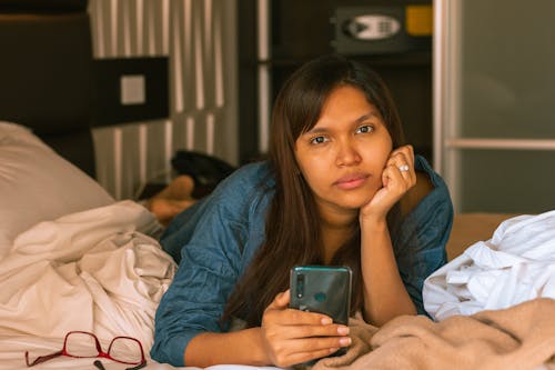 Free A Woman in Denim Shirt Lying on the Bed while Holding Her Mobile Phone Stock Photo