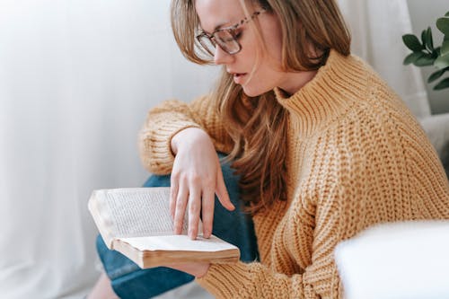 Free Crop female in casual clothes sitting near white curtain and reading interesting book while spending time at home Stock Photo