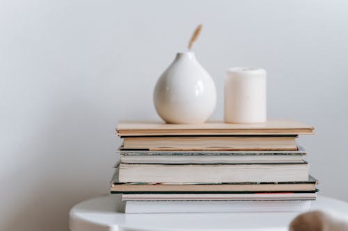Composition of white vase and candle with pile of books on table on white background