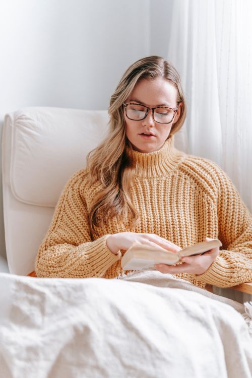 Free Charming young woman in glasses and sweater sitting in comfortable armchair with white plaid and reading book in light white room Stock Photo