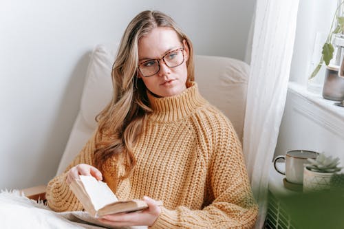 Free Calm thoughtful female in cozy sweater and looking away and while resting in cozy armchair with book Stock Photo
