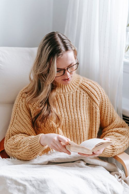 Free Tender female in warm sweater and eyeglasses reading book while sitting in armchair with plaid and spending weekend at home Stock Photo