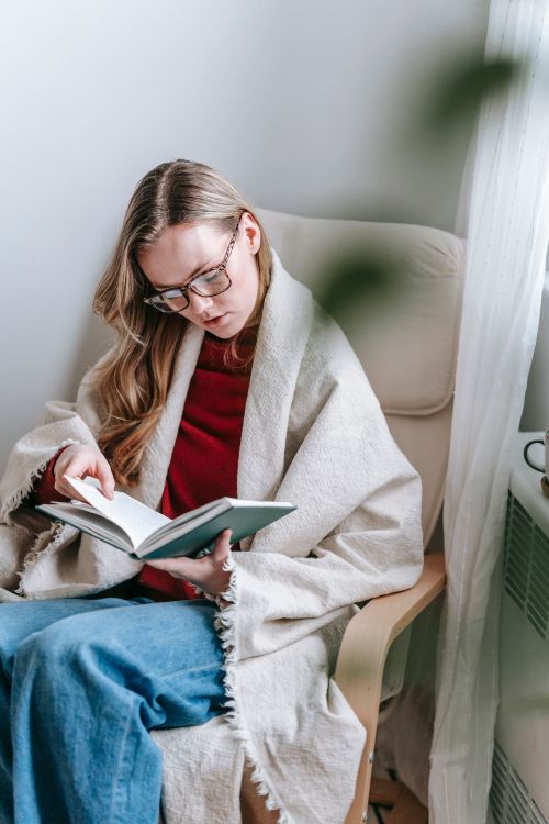 Free Pensive young woman in casual outfit and plaid reading book while sitting in armchair near curtain in bright house Stock Photo