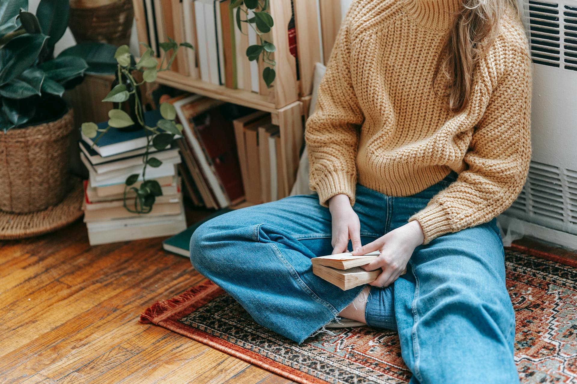 Crop unrecognizable female in stylish warm sweater and jeans sitting on floor at home while reading interesting novel during weekend