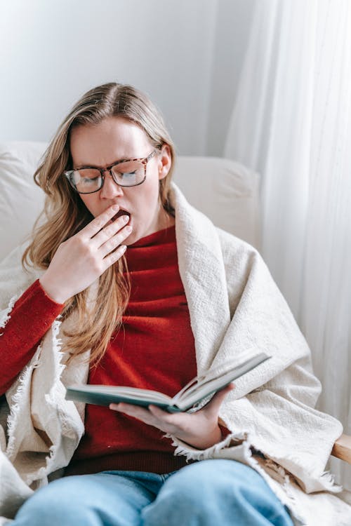 Free Exhausted young woman yawning while reading textbook in armchair Stock Photo