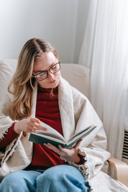 Intelligent young female with long blond hair in warm clothes and eyeglasses sitting on comfortable armchair and reading interesting book during weekend at home