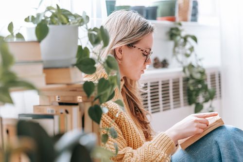 Free Side view of focused young lady with long blond hair in knitted sweater and eyeglasses reading novel sitting on floor near bookshelf in light apartment Stock Photo