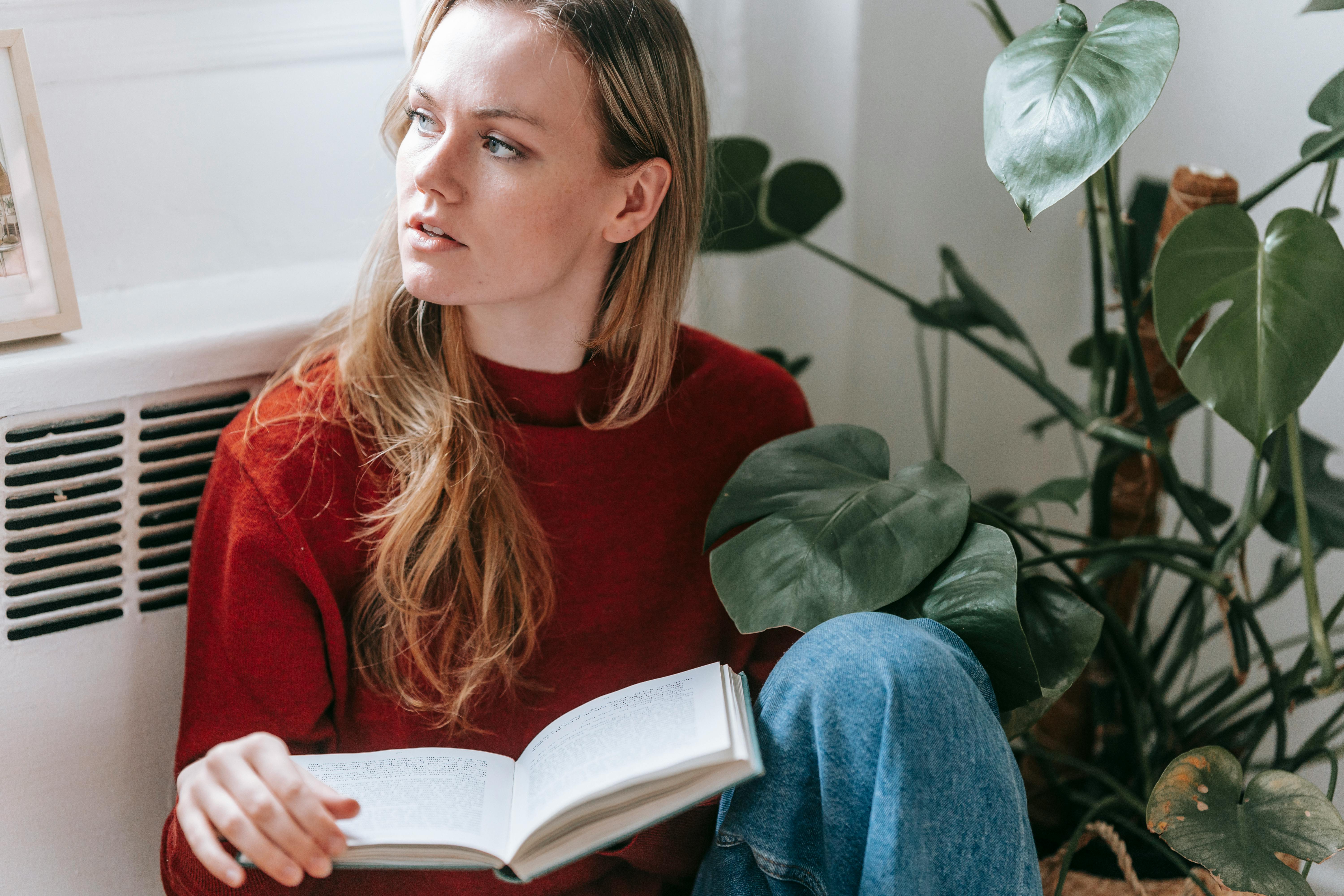 Free Crop thoughtful young woman with long blond hair sitting on floor with book in hand near potted plant and looking away dreamily Stock Photo