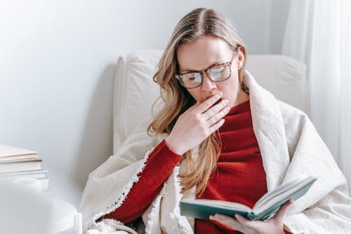 Free Tired young lady with long blond hair in warm clothes and eyeglasses yawning and covering mouth with hand while reading book sitting in armchair at home Stock Photo