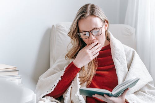 Tired young lady with long blond hair in warm clothes and eyeglasses yawning and covering mouth with hand while reading book sitting in armchair at home